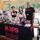 Three individuals stand behind a promotional table for HIPS, an organization called “Honoring Individual Power & Strength.” They are all smiling. The person on the left is wearing black shirt, they have short black hair and wear glasses, they are holding an informational pamphlet with one hand while putting their thumb up with the other; on the middle, a person is holding a small HIPS flag with both hands, they are wearing a white jacket and a headband on their dark medium-size hair; the person on the right wears a shirt that reads “Be nice to sex workers” next to a smiley face, they are wearing a grey cap with a text on it. The table is covered with a black cloth featuring the HIPS logo in pink and the slogan “HARM REDUCTION SAVES LIVES.” It’s decorated with brochures, flyers, candy, buttons, stickers, and other branded materials. Before them are display boards with additional visuals and information about HIPS and its mission.