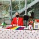 A person wearing a bright red blazer and a black face mask is seated behind a table. The table is covered with a white cloth decorated with rainbow patterns and is filled with pride-themed items, including rainbow-colored beads, small pride flags, informational pamphlets, a QR code sign that reads “Crushing Colonialism” on top, and a hand sanitizer. Behind the person is a glass wall revealing a staircase and an outdoor view with parked cars and greenery.