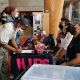 A group of people gathers around a table. The table is covered with a black cloth featuring a word “HIPS” large pink letters. Three individuals sit behind the table, two are engaging with attendees while one of them looks directly to the camera. One person stands while filling out a form on a clipboard, and another stands nearby observing. The table is decorated with colorful items, including flowers, candy, and informational materials. All visible individuals are wearing face masks. The background includes a large wooden column, glass windows, and a hallway.