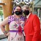 Jen, standing on the left, wears a black face mask and a vibrant dress adorned with geometric and symbolic patterns, including triangles, arrows, and handwritten script designs. Their outfit features a striking red fringe necklace. They have purple hair and are standing closely beside another individual with gray hair, glasses, and a bright red suit jacket over a white shirt and black tie. Behind them is a decorative banner with black, white, and gold geometric designs, and a wooden architectural structure of the venue is visible to the side.