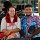 9a, a person with long red hair and round glasses, is seated closely beside another individual. She is wearing a white t-shirt with red and black text that reads “[...] Supreme LAW OF THE LAND” and is smiling warmly. Next to her is Mohammed Zahif, with short curly hair, dressed in a brightly colored shirt with a blue and red abstract pattern, and carrying a small black crossbody bag. Both appear relaxed and cheerful. Behind them, other attendees are seated and working at a table with electronic equipment. Large windows in the background let in natural light and offer a view of the outdoors.