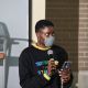 A person is speaking into a microphone while holding a smartphone and looking straight ahead. They are wearing a face mask and a black sweatshirt with bold, colorful text that reads “I CAN’T EVEN THINK STRAIGHT” The setting is indoors, with a brick wall and a chair visible in the background.