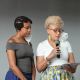 Two individuals stand side by side against a plain gray background. The person on the left wears a gray off-the-shoulder top and blue bottoms, with braided hair and hoop earrings. The person on the right wears a white top and a colorful floral-patterned skirt, holding a microphone and a sheet of paper, suggesting they are reading aloud. Both have face masks hanging from one ear.