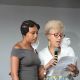 Two individuals stand close together against a plain gray background, engaged in a shared activity. One person holds a microphone and a sheet of paper, reading aloud. Both have face masks pulled down below their chins. The person on the left wears a gray off-the-shoulder, hoop earrings and intricately braided hair. The person on the right has short, curly blond hair, glasses, a white top, and a vibrant, colorful skirt.
