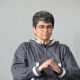 A person with short, curly, salt-and-pepper hair is seated indoors against a plain gray background. They wear glasses and a gray windbreaker-style jacket with white and green accents. Their hands are clasped together in front of them, suggesting a thoughtful or expressive gesture. The lighting is soft and even, indicating a calm, focused setting.