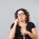 A sign interpreter with curly hair and large black glasses is shown against a plain light gray background. They wear a black top and vibrant accessories, including colorful earrings, a necklace, and a bracelet. The individual is gesturing with one hand, pointing near their eye. The handmade look of the accessories adds a unique and artistic flair to their appearance.