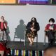 A panel discussion on a stage with three seated women. From left to right: Oswin is speaking through the microphone on her left hand, they have short black hair and glasses, are wearing a red top, black vest, and checkered pants; Johnnie is seated by their side, they have long dark hair and wear a black mask, a black blazer over a floral dress; Daniele is at the end of the row looking at her phone on her lap, she has short curly hair and is wearing a red top and black skirt. Three people appear in front of the stage. The stage edge is decorated with rainbow-striped banners. The backdrop is a plain light gray wall. It’s possible to see the bottom of three flags above the panelists.