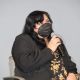 Johnnie sits on a stage chair, speaking into a handheld microphone, they are looking up. They wear a black mask covering her nose and mouth, but appear to be smiling, they have shoulder-length dark hair. Their outfit includes a black blazer over a floral-patterned dress in warm tones. They are seated in a neutral gray background, it’s possible to see Daniele’s arm and curly dark hair on their right left side.