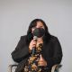 Johnnie sits on a stage chair, speaking into a handheld microphone. They wear a black mask covering her nose and mouth, but appear to be smiling, they have shoulder-length dark hair. Their outfit includes a black blazer over a floral-patterned dress in warm tones. They are seated in a neutral gray background setting with two other chairs partially visible on either side in the scene.