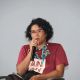 Daniele is speaking into a handheld microphone while her eyes are turned to her front. She has short curly black hair, wears glasses, and is dressed in a maroon short-sleeve shirt with a printed design on the front and a blue-green necklace. She sits in a light gray chair with an armrest, and a cellphone rests on her lap. The background is a plain light gray wall.