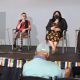 A panel discussion is taking place on a small stage. Four people sit in a row of chairs, each holding a microphone. From left to right: a sign interpreter with black glasses and curly hair wearing a black top and shorts signs to the public; Oswin is speaking while holding the microphone with their right hand and a cellphone with the other, she has short black hair and glasses, is wearing a red top, black vest, and checkered pants; Johnnie is seated by their side listening, they have long dark hair and wear a black mask, a black blazer over a floral dress; Daniele is at the end of the row looking to Oswin, she has short curly hair and is wearing a red top and black skirt. A person in the foreground sits with their back to the camera, watching the panel. The stage edge is decorated with rainbow-striped banners. The backdrop is a plain light gray wall.
