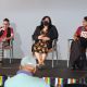 A panel discussion on a stage with three seated women. From left to right: Oswin is looking at her phone while speaking through the microphone on her right hand, they have short black hair and glasses, are wearing a red top, black vest, and checkered pants; Johnnie is seated by their side, they have long dark hair and wear a black mask, a black blazer over a floral dress; Daniele is at the end of the row looking at Oswin, she has short curly hair and is wearing a red top and black skirt. Four people’s heads are seen at the bottom of the image in front of the stage. The stage edge is decorated with rainbow-striped banners. The backdrop is a plain light gray wall.