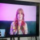 A large TV screen shows a video call. On the main screen, Mandy is speaking into the camera, she has long, wavy ginger hair dyed pink and green with bangs, she is wearing a colorful patterned shirt, and sits against a purple-pink background. She has bold earrings and appears cheerful. In the lower-left corner, a smaller inset video shows another person with dark hair in a black shirt, signing to the public.