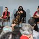 A panel discussion on a stage with three seated women. From left to right: Oswin is looking directly to the public, they have short black hair and glasses, are wearing a red top, black vest, and checkered pants; Johnnie is seated by their side speaking with the microphone on their right hand and gesticulating with the other, they have long dark hair and wear a black mask, a black blazer over a floral dress; Daniele is at the end of the row looking at Johnnie, she has short curly hair and is wearing a red top and black skirt. Four people’s heads are seen at the bottom of the image in front of the stage. The stage edge is decorated with rainbow-striped banners. The backdrop is a plain light gray wall.