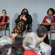 A panel discussion on a stage with three seated women. From left to right: Oswin is looking directly to the public, they have short black hair and glasses, are wearing a red top, black vest, and checkered pants; Johnnie is seated by their side speaking with the microphone on their right hand and gesticulating with the other, they have long dark hair and wear a black mask, a black blazer over a floral dress; Daniele is at the end of the row looking at the crowd, she has short curly hair and is wearing a red top and black skirt. Four people’s heads are seen at the bottom of the image in front of the stage. The stage edge is decorated with rainbow-striped banners. The backdrop is a plain light gray wall.
