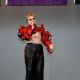 Ritni Tears is speaking on the microphone with an open red jacket that reveals a toned midsection, the other hand is gesticulating with spread fingers. He wears black pants with red stitching and a silver belt, and has short blond hair with heavy makeup around the eyes with white contact lenses. The bottom of banners are seen behind him.