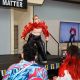 Ritni Tears is performing on the stage striking a dramatic pose with an open red jacket that reveals a toned midsection. He wears black pants with red stitching and a silver belt, and has short blond hair with heavy makeup around the eyes with white contact lenses. The performer is mid-dance, with arms raised and fingers spread, looking upward. A seated observer sits to the left on the stage, smiling, wearing a dark hoodie and khaki pants. The stage edge is decorated with a LGBTQIA+ striped fringe. A large gray wall forms the backdrop, and a couple of flags hang above the stage.