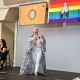 Tony Enos is performing on a black stage. He wears a shiny, sparkling silver sequined jacket, matching white pants with small sparkly embellishments, including a long, patterned cape. He has a white headband and is holding a microphone in one hand while gesturing with the other, as if addressing the audience. Behind him, three banners hang from the ceiling: an orange flag with a seal, a rainbow flag with a central symbol, and a black banner reading “BLACK LIVES MATTER.” An interpreter sits to the left on a chair, signing the performance to the performance. The stage has a plain gray backdrop, and the front edge is decorated with a LGBTQIA+ striped fringe.