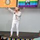 Tony Enos is performing on the stage, singing into a microphone. He wears a sparkling silver sequined jacket, matching white pants with small sparkly embellishments, and a white headband. He is mid-gesture with one arm raised and the other hand holding the microphone. Behind him are banners hanging from the ceiling, including a rainbow flag and another orange flag with a circular emblem. An interpreter sits off to the left on a chair, signing the performance to the public. The stage is indoors with a gray wall and a small LGBTQIA+ fringe along the front edge.