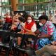 A group of people seated in a row of black folding chairs inside a bright venue. Most are wearing face masks. In the foreground, a person in a turquoise and white patterned outfit sits next to a person in a red and white top with glasses and a white mask, both looking at their phones; a woman in a red scarf sits beside them, also masked. Several attendees raise one hand waving their cellphone flashlights, while others look toward the stage or camera. A few bags and personal items rest on the chairs. The background shows large windows with a street view and some standing photographers or videographers. Overall, it looks like a casual, inclusive event with a diverse audience.