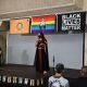 Yaffa is standing on a stage and speaking into a microphone. They are wearing a long black dress with red decorative patterns and pink sandals, and have a black face mask on. They hold a small notebook in one hand. Behind them, three banners are hanging: a colorful pride flag, a flag with an emblem, and a black banner that reads “BLACK LIVES MATTER.” To the left, the sign interpreter sits on a chair, wearing a black dress and shoes. The audience is seated, and some people are visible in the foreground. The stage is simple with a plain gray backdrop, and there is a decorative rainbow-striped border along the front of the stage.