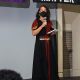 Yaffa is standing on a stage, wearing a long black dress with red decorative patterns and pink sandals, holding a microphone and a small notepad. They are wearing a black face mask. Behind them are banners, including a rainbow flag and a black banner that reads "BLACK LIVES MATTER." The setting appears to be an indoor event with a neutral gray wall and brick pillars in the background.