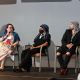 Three panelists sit on stage during a discussion. Jen, wearing a colorful dress and holding a microphone, speaks while looking at the others. Rivolta, wearing a blue-and-white striped headscarf and black mask, sits with hands folded in their lap and looks towards the speaker. Tony, dressed in black with a patterned black-and-white scarf and red shoes, looks toward the speaker. Audience members are partly visible in the foreground.