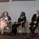 A panel discussion is taking place on a small stage. Three people sit in a row of chairs. From left to right: Jen is speaking into a handheld microphone while smiling, a they have short dyed purple hair, black glasses and are wearing a long dress with geometric designs and a blue scarf; Rivolta Sata is next in line looking at Jen, they are wearing black pants, t-shirt and shoes, a white necklace, a black and blue striped headscarf covering their hair and a black mask; Tony Enos sits at the end of the row looking and smiling at Jen, he wears black pants ripped on the knees, a black jacket and t-shirt, a checkered black and white headscarf. A mobility scooter is partially visible on the far left of the image.