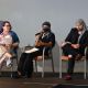 A panel discussion is taking place on a small stage. Three people sit in a row of chairs. From left to right: Jen is holding sheets of paper while looking at Rivolta Sata, they have short dyed purple hair, black glasses and are wearing a long dress with a pattern and a blue shawl; Rivolta Sata is next in line, they hold a microphone with one hand and gesticulates with the other, they are wearing black pants, t-shirt and shoes, a white necklace, a black and blue striped headscarf covering their hair and a black mask; Tony Enos sits at the end of the row crossing his legs, he wears black pants ripped on the knees, a black jacket and t-shirt, a checkered black and white headscarf.