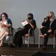 Three panelists sit on stage during an event. On the left, Jen is wearing a colorful dress, looking at a paper in their hand. Rivolta, wearing a headscarf and mask, also holds a paper while seated. Tony, dressed in black with a patterned scarf, speaks into a microphone. Audience members are partially visible in the foreground.