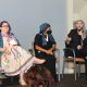 Three panelists sit on stage during a discussion. From left to right: Jen, with purple hair, glasses, a colorful patterned dress, and a blue shawl, smiling with legs crossed beside a mobility scooter draped with an orange flag; Rivolta, dressed in black with a blue-and-white striped headscarf and black face mask, sitting with hands folded looking at Tony; and Tony, in a black suit jacket, ripped black jeans, red shoes, and a black-and-white patterned headscarf, speaking into a microphone while gesturing with his handand smiling. Audience members are visible in the foreground.