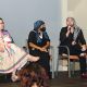 Three panelists are seated on stage during a discussion. From left to right: Jen, with purple hair, glasses, a colorful dress, and a blue shawl, looking toward the others; Rivolta, dressed in black with a blue-and-black striped headscarf and a black face mask, sitting with hands folded; and Tony, in a black suit jacket, ripped black jeans, red shoes, and a black-and-white patterned headscarf, speaking into a microphone. A mobility scooter draped with an orange flag is partly visible on the far left, and audience members are seen in the foreground.