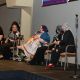 Five people sit on stage during a panel discussion. From left to right: a sign language interpreter dressed in black; Lady Shug, a drag performer in a long black gown with bold makeup, leans over the chair’s armrest; Jen, with purple hair, glasses, a colorful dress, and a blue shawl, speaking into a microphone, by their side there’s a yellow mobility scooter; Rivolta, in black clothing with a blue-and-white striped headscarf and black face mask, looks at Jen; and Tony, in a black suit jacket, ripped black jeans, red shoes, and a black-and-white patterned headscarf, looking toward the others. A large screen on the right shows a live feed of the panel.