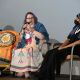 Two people are seated on a stage during a panel discussion. On the left, Jen is speaking into a microphone. They are wearing a colorful dress with geometric designs and a blue shawl, they have purple hair and wear glasses. Next to them is a yellow mobility scooter draped with an orange flag featuring a circular seal with a star and text, along with several small flags. To the right, Rivolta is dressed in black clothing with a blue-and-white striped headscarf and a black face mask listening attentively. Part of Tony, dressed in black, is partially visible on the far right.