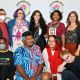 A group of nine individuals pose together in front of a white backdrop that reads “DECOLONIZED BEATZ INDIGENOUS WORLD PRIDE” Standing, from left to right: Oswin wears a red t-shirt, a black vest, checkered pants and holds a pride flag and a white mask; Jen stands next, dressed in a long dress with geometric designs, a blue shawl and a fringe necklace, they have short purple hair and wear glasses. Fabiana follows, wearing a black dress with a toucan print with a relaxed and cheerful expression, she has long black wavy hair. Daniele stands beside her, dressed in a red t-shirt, wearing a blue feather necklace and smiling softly. The fifth person stands with a gentle smile, she’s wearing a black top with geometrical brown and white geometrical shapes, a black cardigan and deep orange pants, she has medium-size brown hair. Kneeling in front, from left to right: a person is wearing a grey mask, black tights and sweater with words that read “I CAN’T EVEN THINK STRAIGHT”, a red bag hangs from their shoulder. Next is Mohammed Zahif in a matching blue set with a red and dark blue pattern, he has a mustache and dark short hair, and is smiling softly with their eyes closed. Next is 9a, wearing a beige oversized shirt, red sweater around their neck, gray shorts, and red cowboy boots, with long red-dyed hair and a camera around their neck. Beside them is Ritni, dressed in black with dramatic makeup and white contact lenses, with their head leaning on their right hand looking at the camera.
