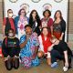 A group of nine individuals pose together in front of a white backdrop that reads “DECOLONIZED BEATZ INDIGENOUS WORLD PRIDE” Standing, from left to right: Oswin wears a red t-shirt, a black vest, checkered pants and holds a pride flag and a white mask; Jen stands next, dressed in a long dress with geometric designs, a blue shawl and a fringe necklace, they have short purple hair and wear glasses. Fabiana follows, wearing a black dress with a toucan print with a relaxed and cheerful expression, she has long black wavy hair. Daniele stands beside her, dressed in a red t-shirt, wearing a blue feather necklace and smiling softly. The fifth person stands with a gentle smile, she’s wearing a black top with geometrical brown and white geometrical shapes, a black cardigan and deep orange pants, she has medium-size brown hair. Kneeling in front, from left to right: a person is wearing a grey mask, black tights and sweater with words that read “I CAN’T EVEN THINK STRAIGHT”, a red bag hangs from their shoulder. Next is Mohammed Zahif in a matching blue set with a red and dark blue pattern, with a lollipop in their mouth, he has a mustache and dark short hair. Next is 9a, wearing a beige oversized shirt, red sweater around their neck, gray shorts, and red cowboy boots, with long red-dyed hair and a camera around their neck. Beside them is Ritni, dressed in black with dramatic makeup and white contact lenses, with their head leaned on their right hand looking down.