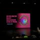 A stage setup in a dark auditorium features a large screen displaying colorful graphics and bold text that reads: “DECOLONIZED BEATZ INDIGENOUS WORLD PRIDE — STARTING SOON!” Next to the text is a circular logo with a globe at the center, surrounded by rainbow-colored beams and the silhouette of a person wearing a feathered headdress. Two individuals sit and talk casually on the stage, while a few audience members are seated in the foreground, waiting for the event to begin. The atmosphere suggests anticipation and excitement for a celebration of Indigenous identity, decolonization, and artistic expression.