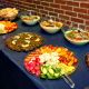 A buffet-style food spread is arranged on a dark blue tablecloth against a brick wall, showcasing a vibrant selection of Mediterranean or Middle Eastern dishes. On the left, a platter holds neatly arranged pita bread alongside sliced cucumbers, red and yellow bell peppers. Next to it, four large glass bowls contain creamy, yogurt-based salads or dips, each garnished with herbs and spices for visual and flavor contrast. A platter of stuffed grape leaves sits nearby, accented with lemon slices. Further along, a colorful vegetable arrangement features radishes, cauliflower florets, cucumbers, cherry tomatoes, and bell pepper strips. On the far right, a platter of grilled meat is garnished with chopped herbs.