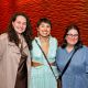 Three women pose to the camera all smiling. The one on the left is wearing a brown satin dress, and a long suede brown jacket, she has long curly brown hair. In the middle, Tatiana stands embracing both, with a ruffled cutout baby blue dress with a V-neck, a crossbody purse, she has short hair and is wearing colorful beaded earrings and white eyeliner. The woman on the left is wearing a button-up blue shirt dress, her hair is middle parted shoulder-length, she’s wearing black glasses, a necklace and a crossbody black purse.