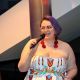 Jen stands holding a microphone, captured mid-speech. They have short, vibrant purple hair and wear a strapless dress with a bold geometric pattern in shades of red, yellow, blue, and black. Large red circular earrings accent their look, adding a pop of color that complements their outfit. Their posture is upright and engaged, with one hand holding the microphone close to their mouth and the other arm holding a paper by their side. Their expressions are animated and confident.
