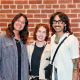 Three individuals stand closely together in front of a red brick wall, smiling warmly as they pose for a photo. On the left, a person wears a black top layered with a checkered jacket and a necklace, their short dark hair styled neatly and their posture relaxed. In the center, a person with long light hair wears a light-colored jacket over a black shirt, with a crossbody black purse. On the right, a person with short curly hair wears a white textured short-sleeve shirt, accessorized with a turquoise necklace, glasses, and a black crossbody bag.
