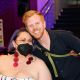 Jen is seated wearing a mask, bold red earrings and a strapless white dress with red and white designs, she has short vibrant purple hair. She also wears a black mask but appears to be smiling under it. A man in a black shirt, wearing a crossbody colorful purse is leaning over her for the picture, he has ginger hair and beard, and is smiling.