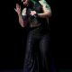 Tony Enos performs on stage wearing a glittery black sleeveless top, flared pants, and a black headpiece. He holds a microphone in his right hand, raised near his mouth, while his left arm extends outward in a sweeping, expressive gesture—fingers splayed as if reaching toward the audience up. His stance is wide and grounded, with visible tattoos on both arms adding to the visual rhythm of his movement.