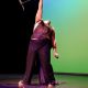 Tony Enos performs on stage in a dramatic pose, dressed in a glittery dark outfit that catches the light. One arm is raised high above his head, gripping a microphone stand, while the other hand holds a microphone near his side. His stance is powerful and theatrical, capturing a climactic moment in the performance. The background glows with a gradient of green and blue lighting, adding intensity and vibrancy to the scene.
