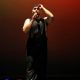 Tony Enos stands center stage under dramatic lighting, wearing a sparkly black sleeveless outfit and a black headpiece. He grips a microphone in his right hand while his left arm is raised above his head, he looks forward with a powerful and focused expression. The background glows with a gradient of red fading into darkness