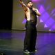 Tony Enos stands mid-performance on stage, wearing a glittery black sleeveless outfit with wide-legged pants and a headpiece. He holds a microphone in his right hand while his left arm is raised in a sweeping gesture, as if emphasizing a powerful lyric or engaging the audience with dramatic flair. His stance is open and expressive, with his torso slightly angled and knees bent, suggesting motion and intensity.