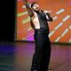 Tony Enos performs on stage in a sparkly black sleeveless outfit with matching pants and a black headpiece. Holding a microphone in his right hand, he raises his left arm in a dramatic, sweeping gesture, fingers extended. His torso twists slightly, emphasizing the upward motion of his arm and the energy of the moment. A tattoo of a bird is visible on the left side of his torso, adding to the visual rhythm of his pose.