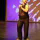 Tony Enos stands mid-performance on stage, wearing a sparkly black sleeveless top, matching wide-legged pants, black shoes, and a black headpiece. He holds a microphone in his left hand, raised near his mouth. His head is turned to his left side, his eyes facing the same direction, and a smile. His right hand is not visible, possibly positioned behind his body or out of frame. His torso leans slightly forward, and his knees are softly bent, suggesting motion and engagement with the audience. Visible tattoos on his left arm.