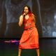 9a stands on stage mid-performance, wearing a bold red tank top and matching skirt. The top features illustrations of two wolves, and the skirt is printed with the words “AHUNSKA” and “MATOLIN” in vertical and horizontal orientations. Their long, straight hair with red highlights flows down their back, and they wear white sandals with red accents. Holding a microphone in their left hand, 9a leans slightly forward, knees bent, with their right arm to their back. Their posture is expressive and grounded.