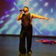 Tony Enos performs on stage wearing a sparkly black sleeveless top, matching pants, black shoes, and a black headpiece. He holds a microphone in his right hand, raised near his mouth singing. His left arm is lifted in a fluid, expressive gesture—fingers extended and palm open. His body leans slightly forward, knees bent, creating a sense of motion and emotional engagement.